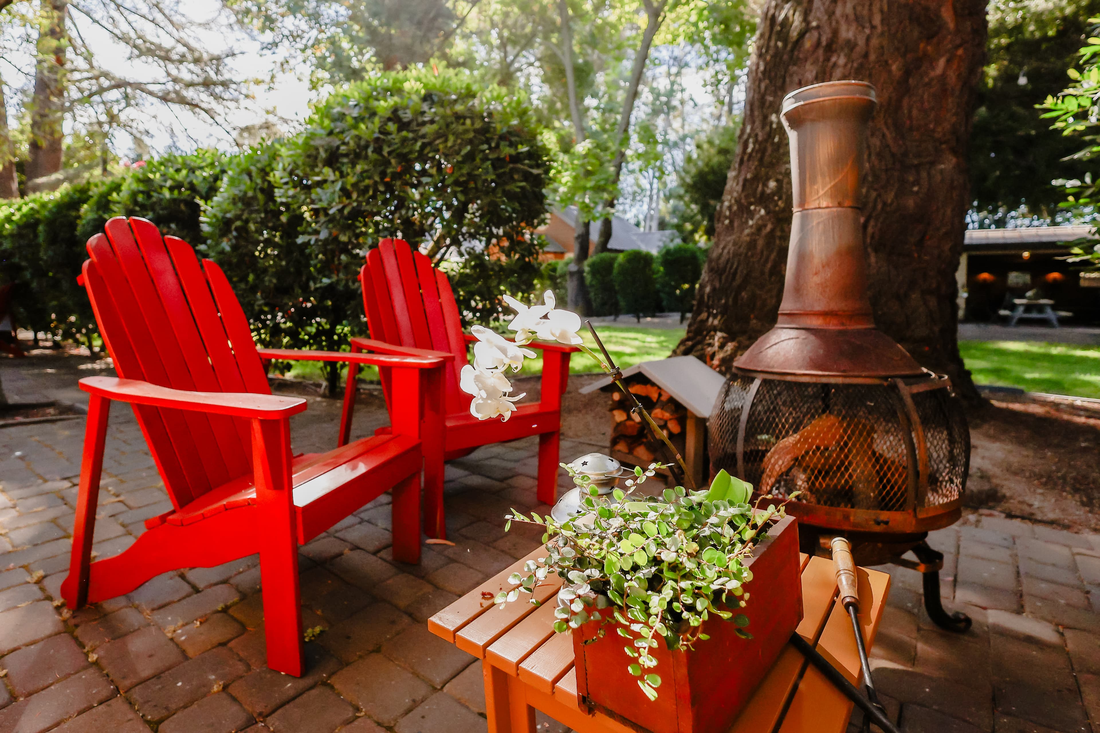 A cozy outdoor seating area featuring two red Adirondack chairs, a potted plant, and a chiminea surrounded by greenery.
