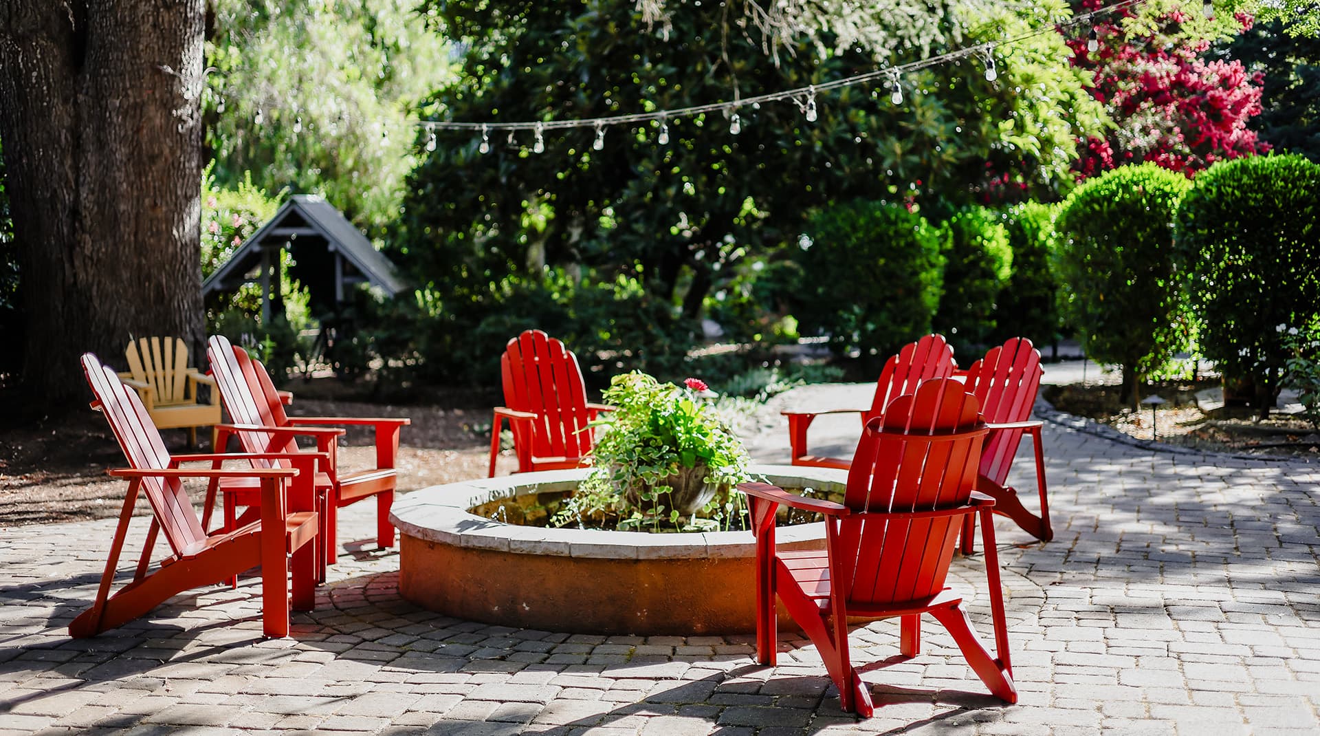 A circular fire pit surrounded by red Adirondack chairs in a lush garden setting.