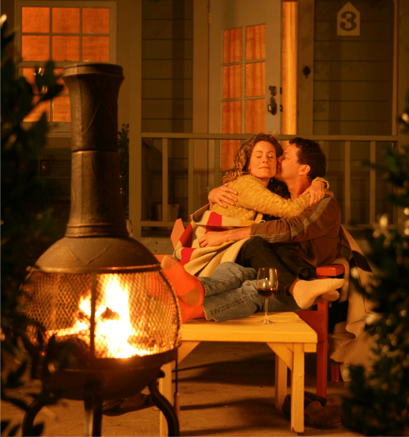 A couple embraces by a cozy outdoor fire, wrapped in a blanket, with a glass of wine nearby.