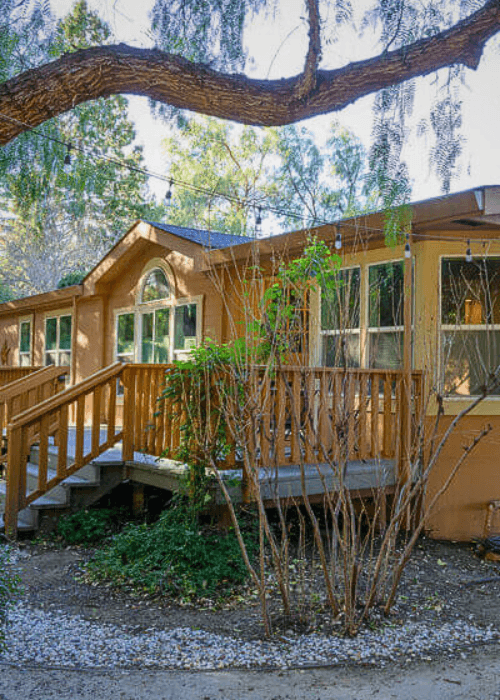A cozy brown house with a wooden porch surrounded by greenery and gravel landscaping.