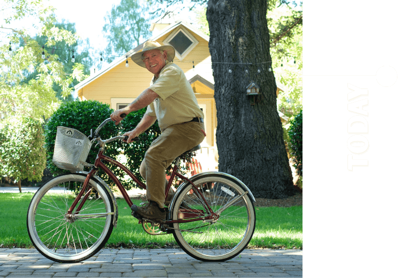 A smiling man in a hat rides a bicycle in a sunny, green yard.