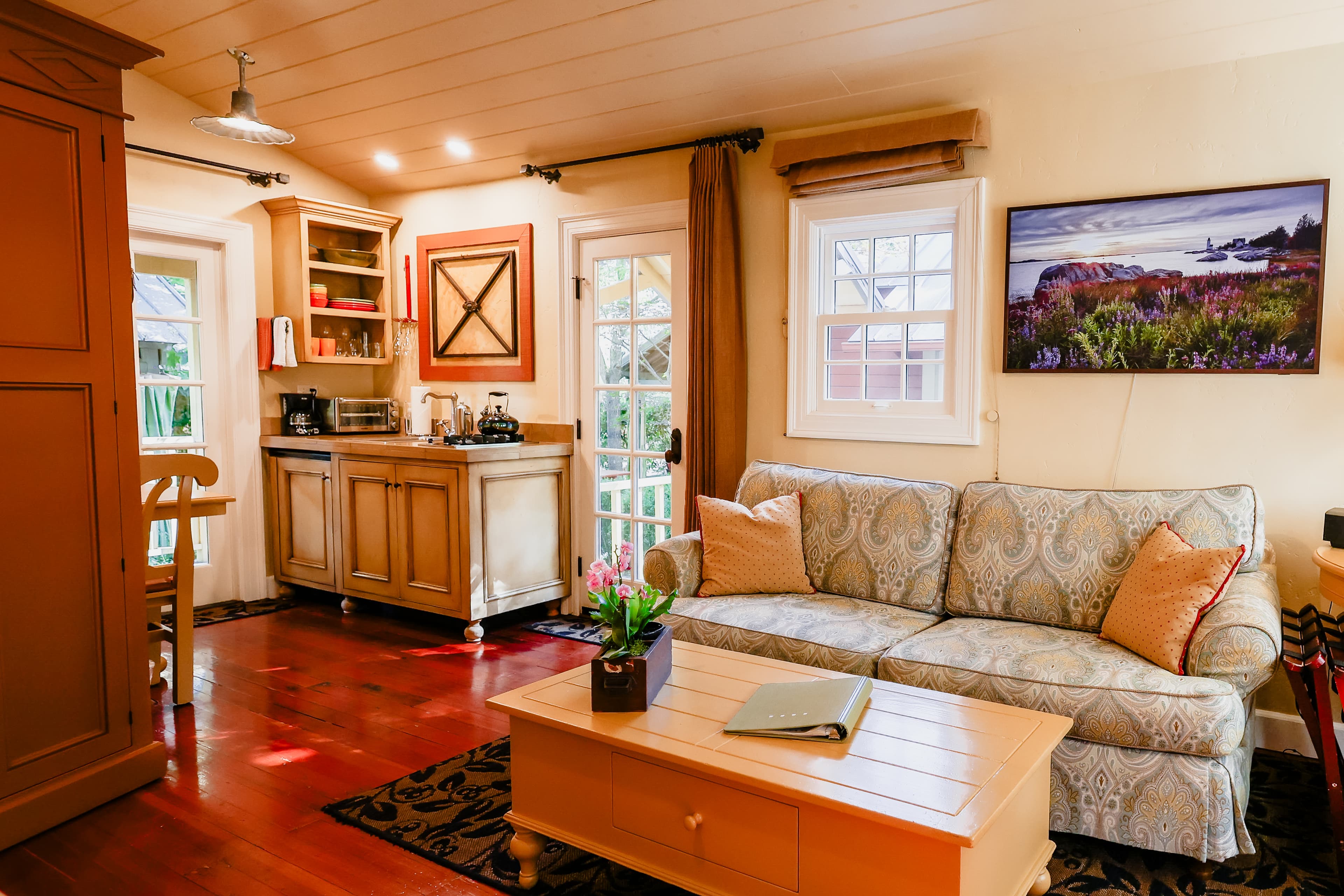 A cozy living space featuring a patterned sofa, coffee table, and kitchenette with natural light from large windows.
