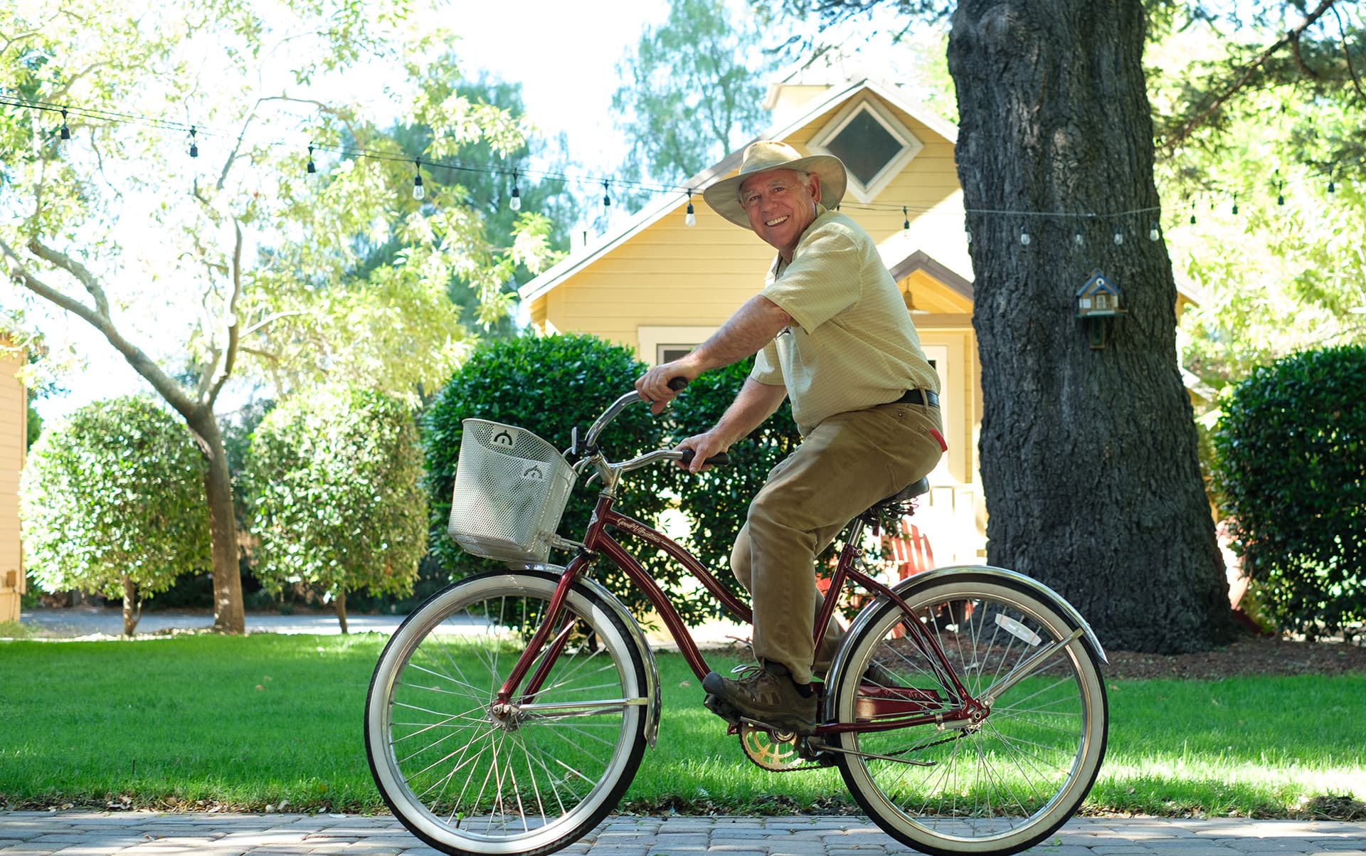 A smiling man rides a bicycle in a sunny garden setting.