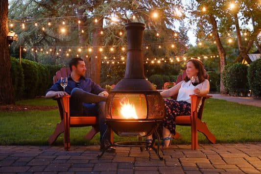 A couple relaxes by a chiminea, enjoying wine in a beautifully lit garden.