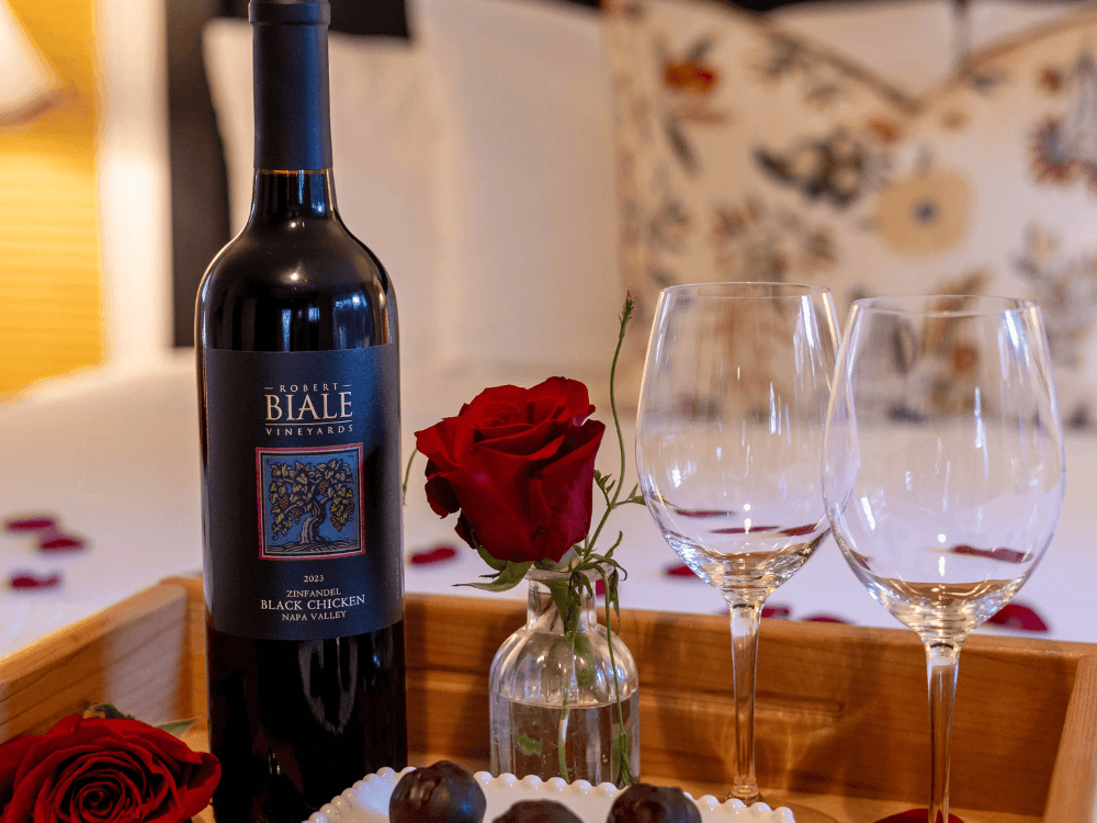 A bottle of red wine is accompanied by two wine glasses, a single rose, and chocolates on a wooden tray.