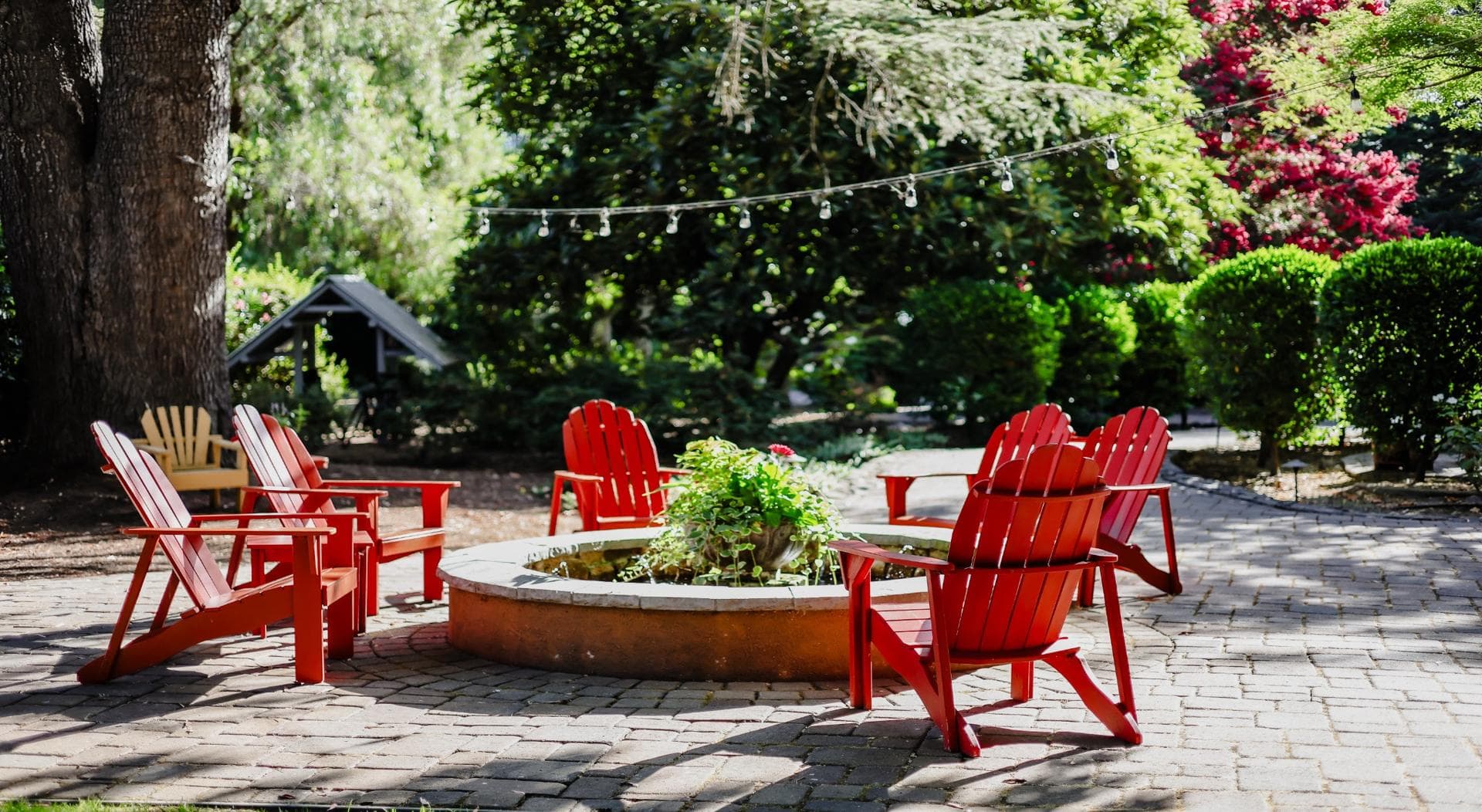 A circle of red Adirondack chairs around a stone fire pit in a green garden.