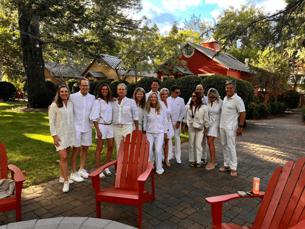 A group of people dressed in white stands together in a lush outdoor setting with red chairs and colorful houses in the background.