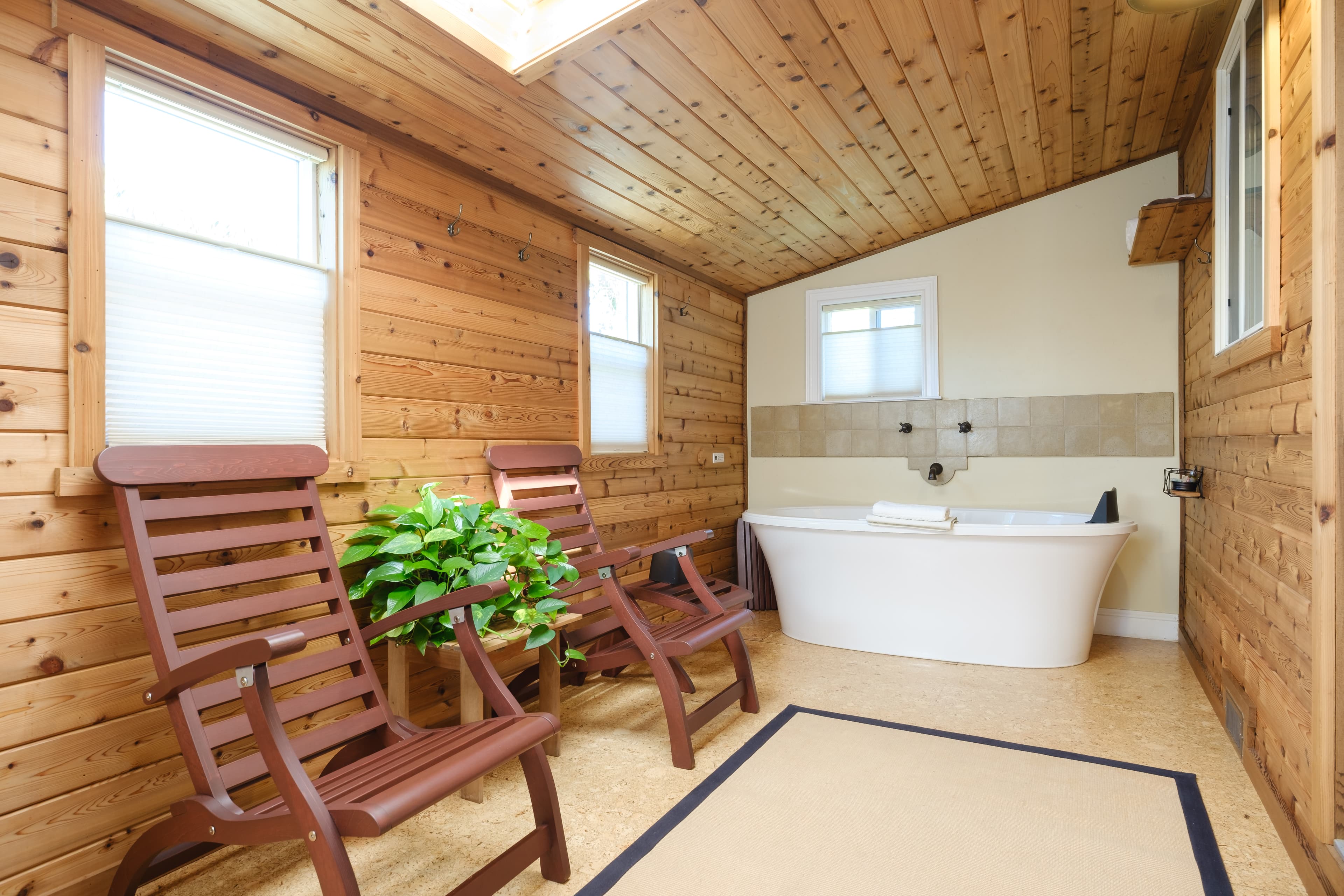 A cozy wooden bathroom featuring two chairs, a large bathtub, and plants by the window.
