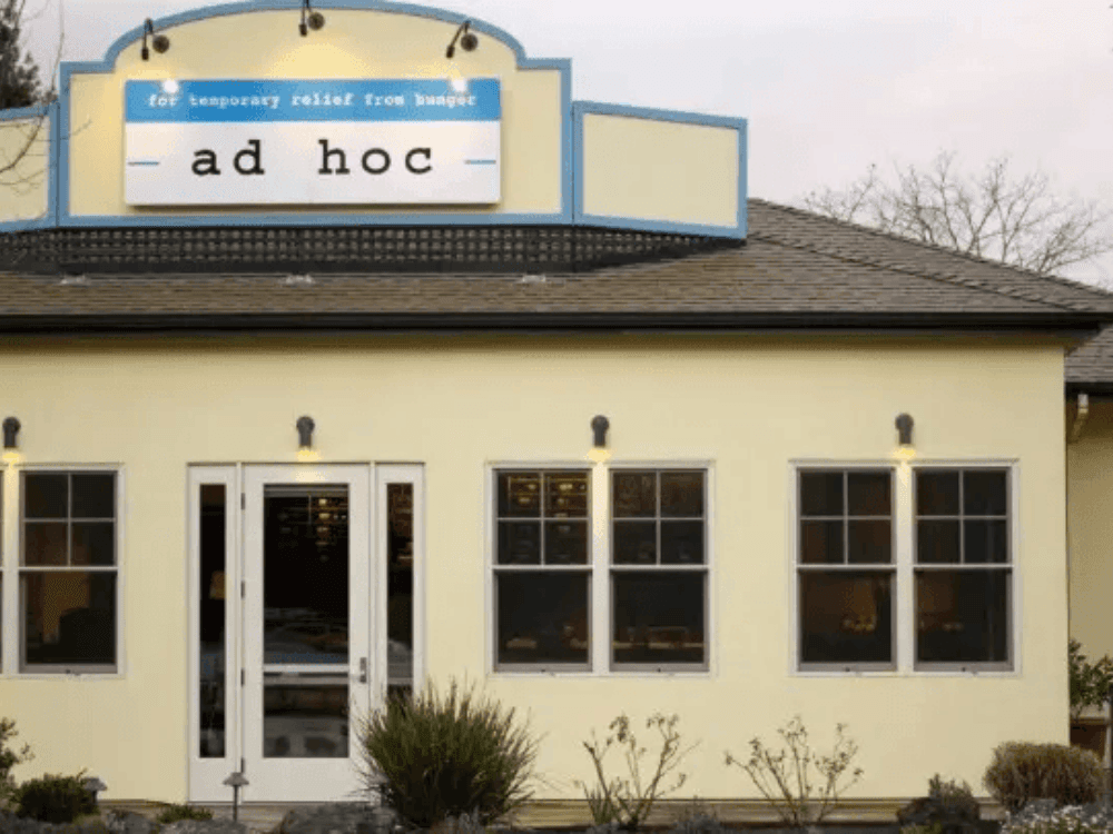 Exterior of a yellow restaurant building with a sign reading "ad hoc" and a tagline about temporary relief from hunger.
