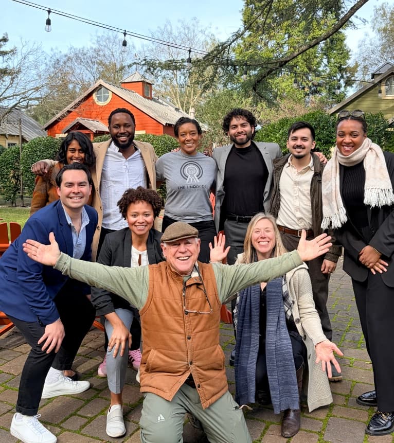 A diverse group of people poses joyfully outdoors in a park-like setting.