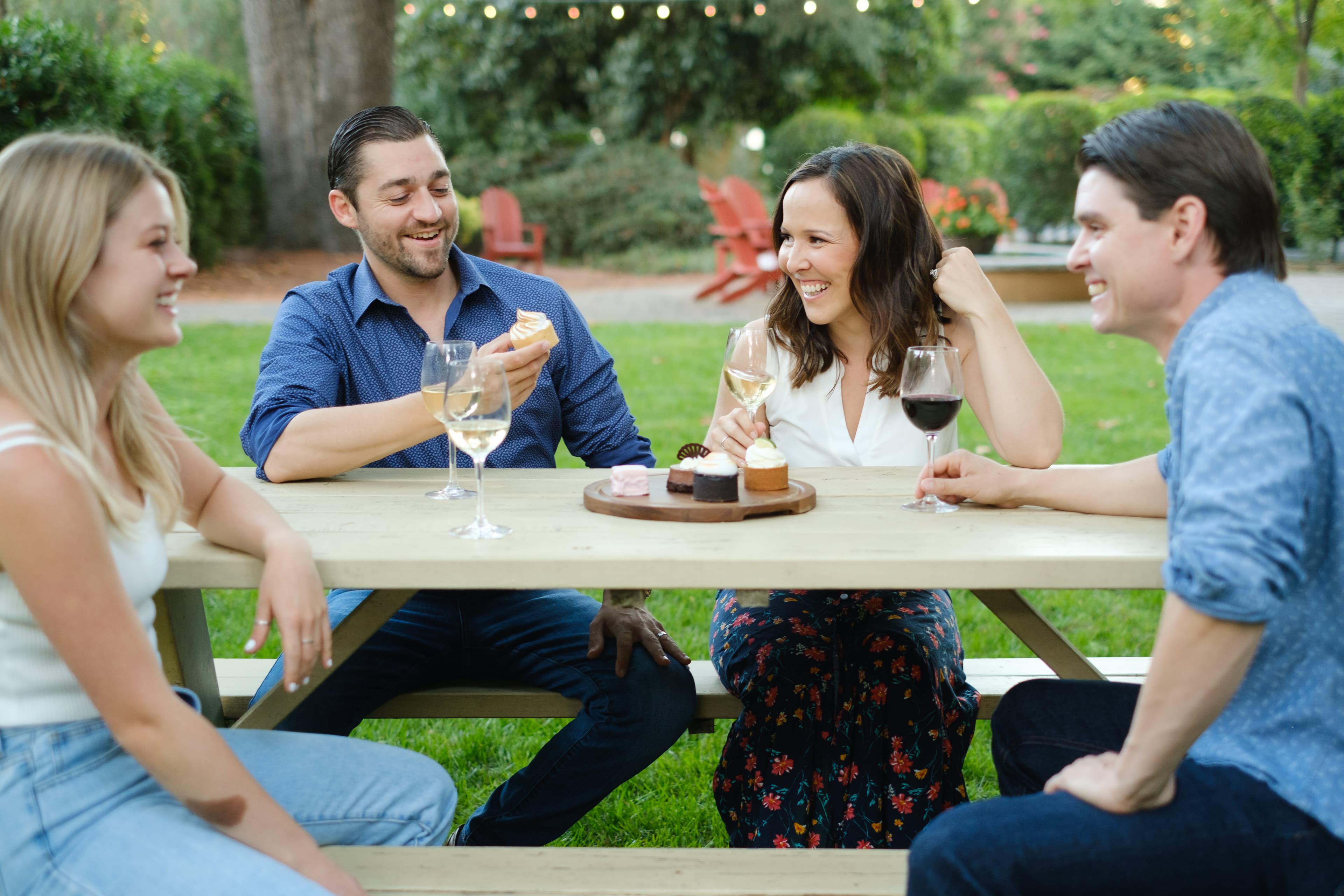 Four friends enjoy dessert and wine at a picnic table in a garden setting.