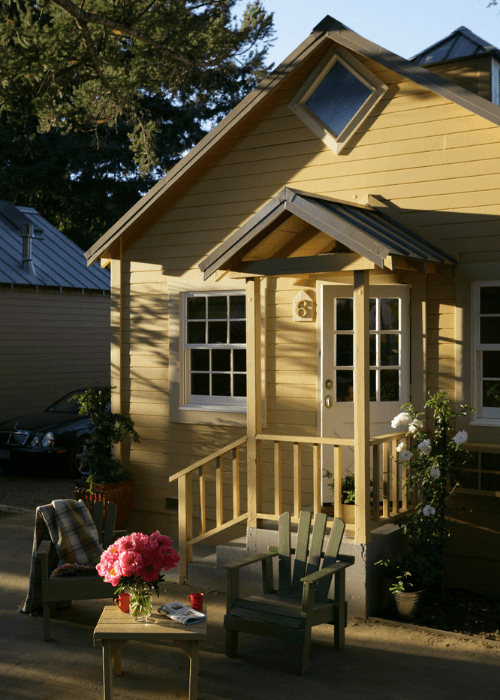 A quaint yellow house with a porch, surrounded by greenery and outdoor furniture.