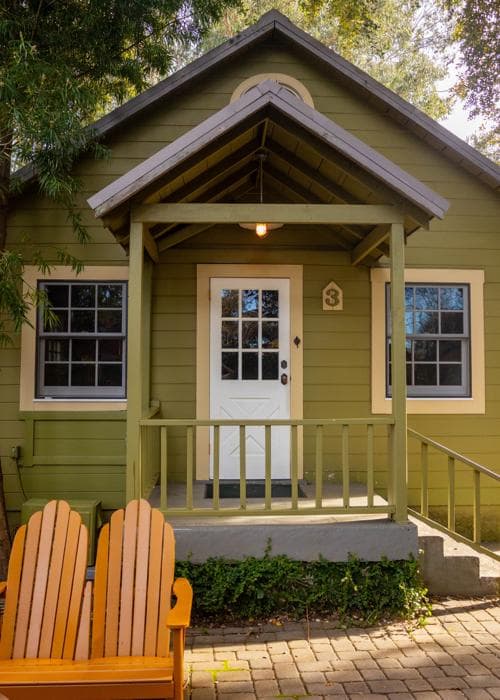 A small green cottage with a porch and two orange chairs in front.