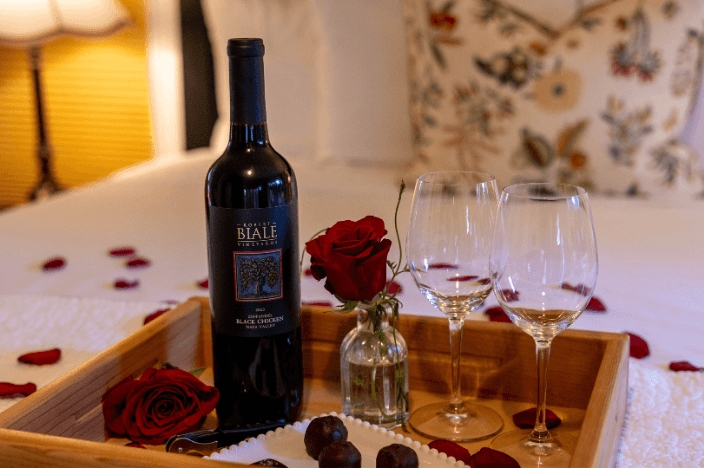 A romantic setup featuring a bottle of wine, two glasses, a rose, and chocolates on a tray, surrounded by flower petals.