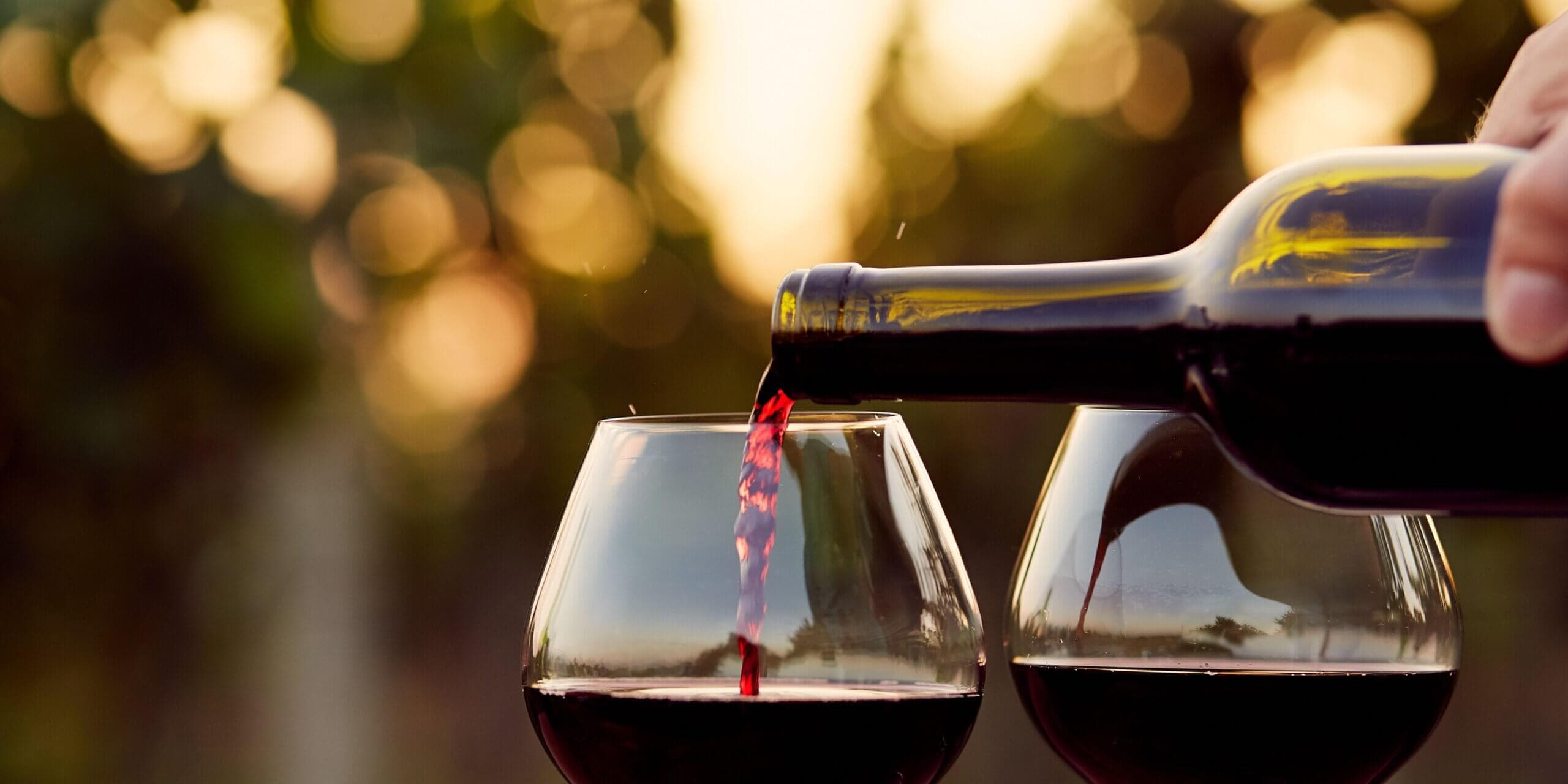 A bottle of red wine is being poured into two glasses against a blurred, sunset vineyard background.
