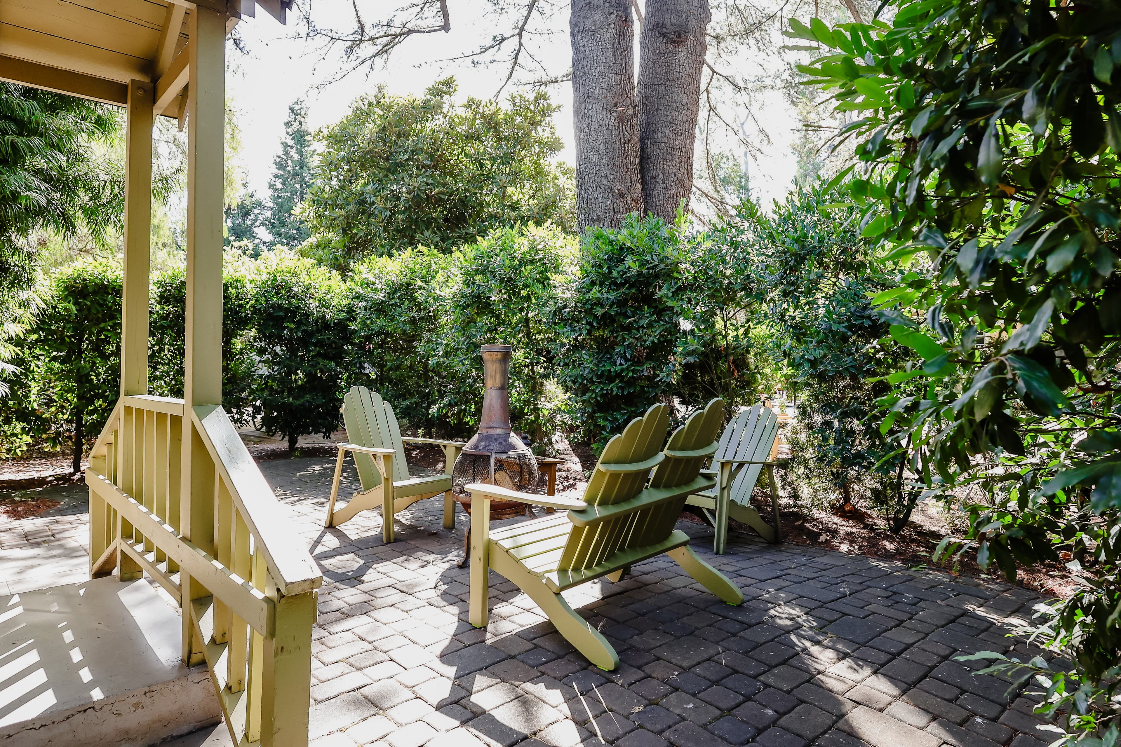 A cozy outdoor seating area with green chairs and a chiminea, surrounded by lush greenery.