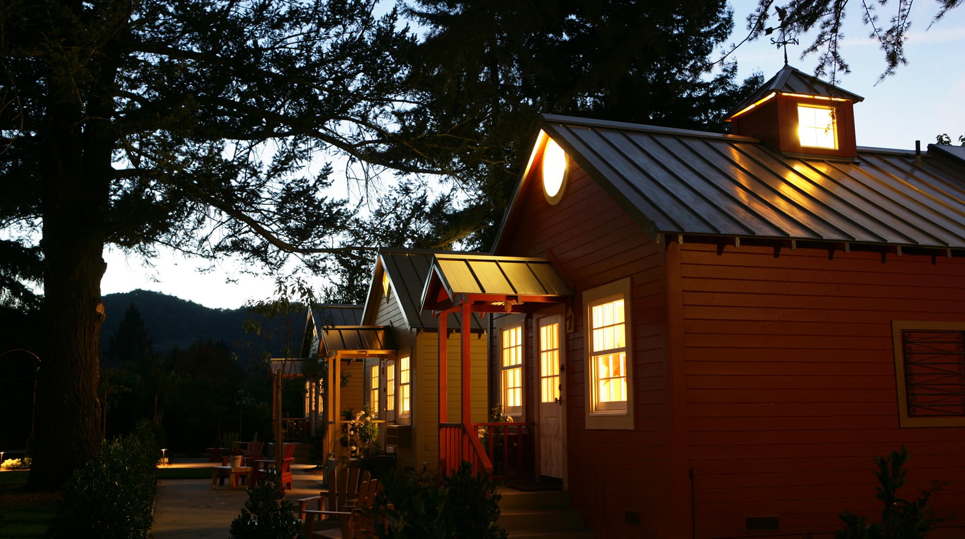 Three cozy cottages illuminated at dusk, surrounded by trees.